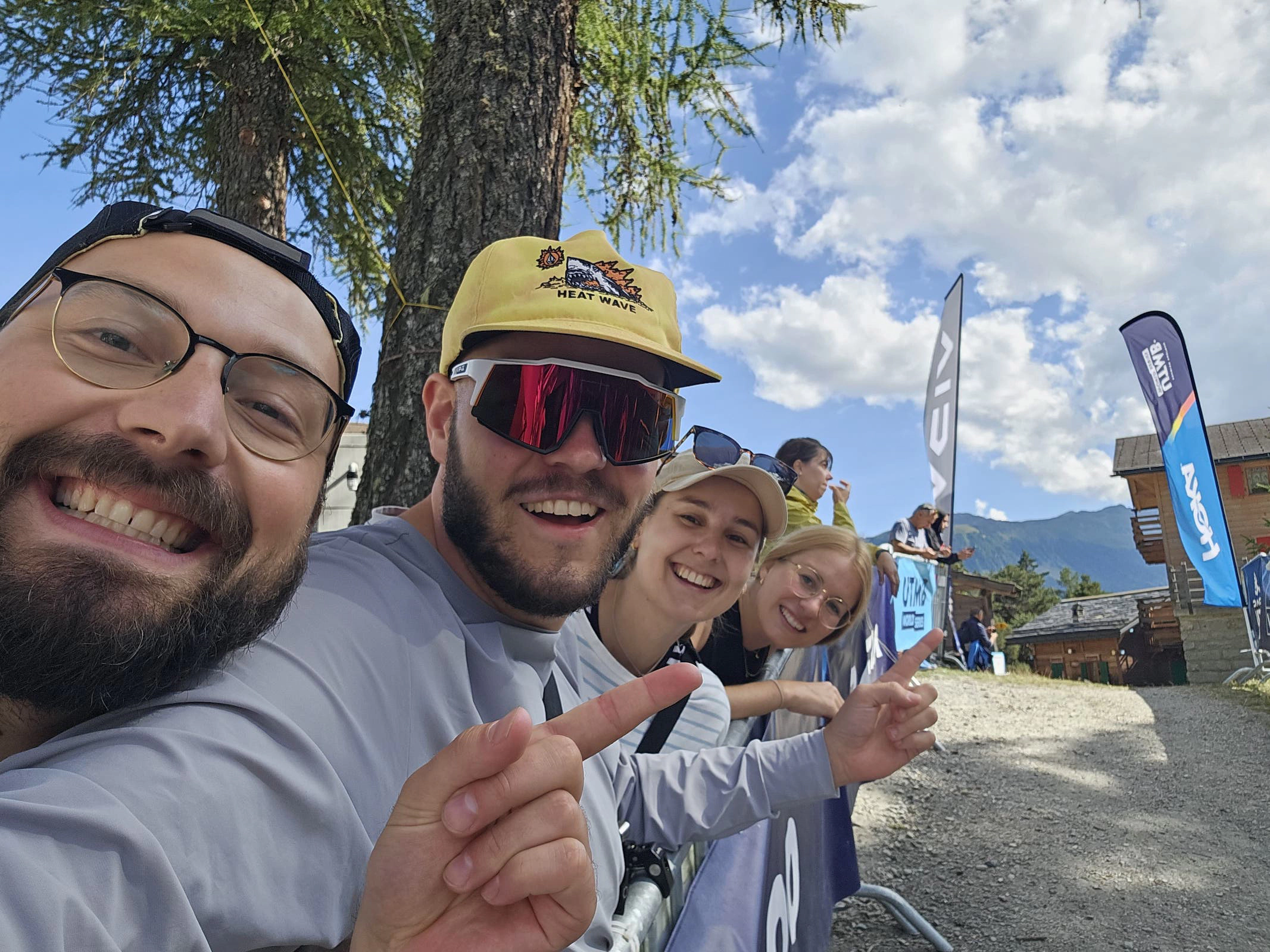 Selfie der Support Crew am Streckenrand hinter Absprerrungen. Im Hintergrund wehen die Fahnen der Sponsoren des UTMB.