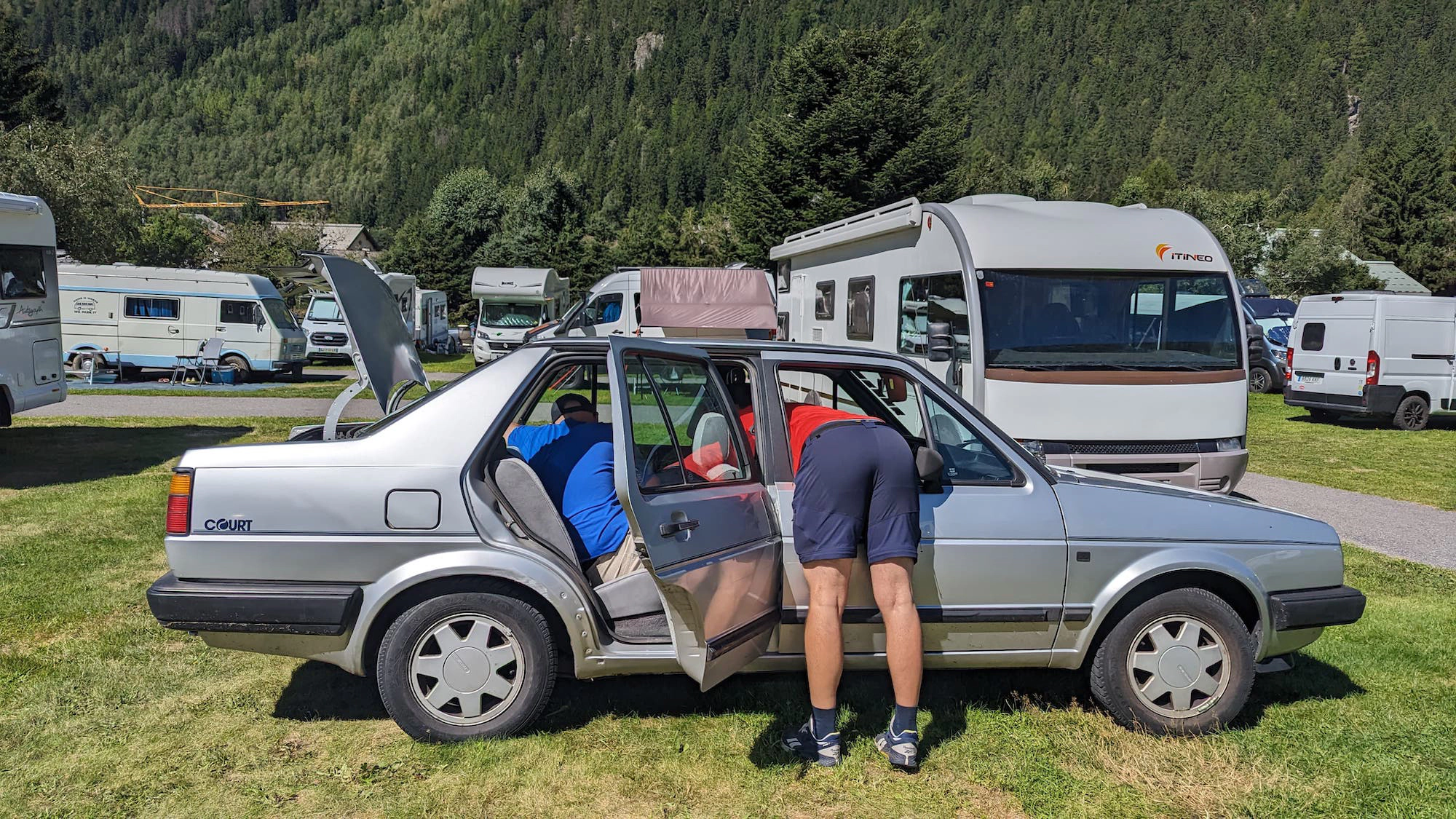 Zwei Männer beladen ein Auto mit geöffnetem Kofferraum. Einer hängt durch die geschlossene Beifahrertür in den Innenraum.