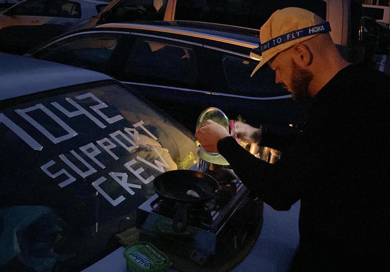 In der Nacht kocht ein Mann auf einem Gaskocher, welcher auf dem geschlossenen Kofferraum eines Autos steht Essen.